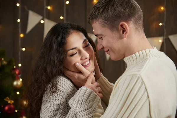 若いカップルは、ライトとダークの木製インテリアのハグします。ロマンチックな夜と愛の概念。年末年始。クリスマスの照明そして装飾。白い服を着て. — ストック写真