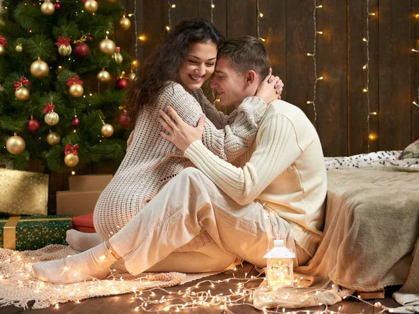 Giovane coppia si siede sul pavimento in legno scuro interno con luci. Serata romantica e concetto d'amore. Vacanza di anno nuovo. Luci e decorazioni natalizie. Vestita di bianco . — Foto Stock
