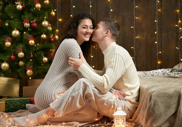 Giovane coppia si siede sul pavimento in legno scuro interno con luci. Serata romantica e concetto d'amore. Vacanza di anno nuovo. Luci e decorazioni natalizie. Vestita di bianco . — Foto Stock