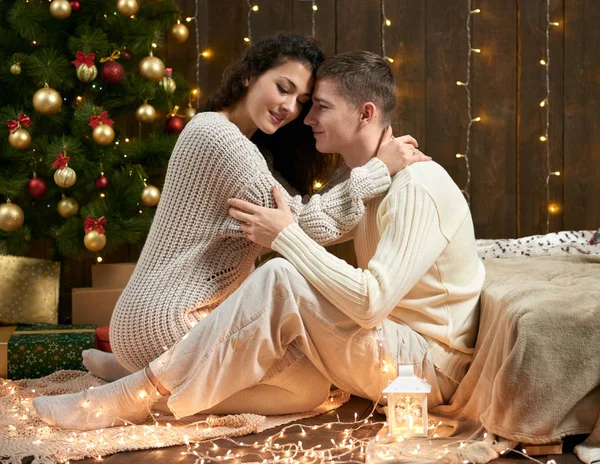 Giovane coppia si siede sul pavimento in legno scuro interno con luci. Serata romantica e concetto d'amore. Vacanza di anno nuovo. Luci e decorazioni natalizie. Vestita di bianco . — Foto Stock