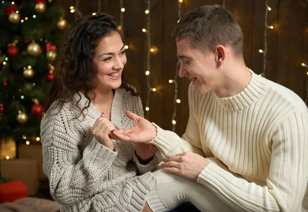 男子给女孩一个订婚戒指, 情侣在圣诞彩灯和装饰, 身着白色, 冷杉树在黑暗的木制背景, 冬季假期概念 — 图库照片