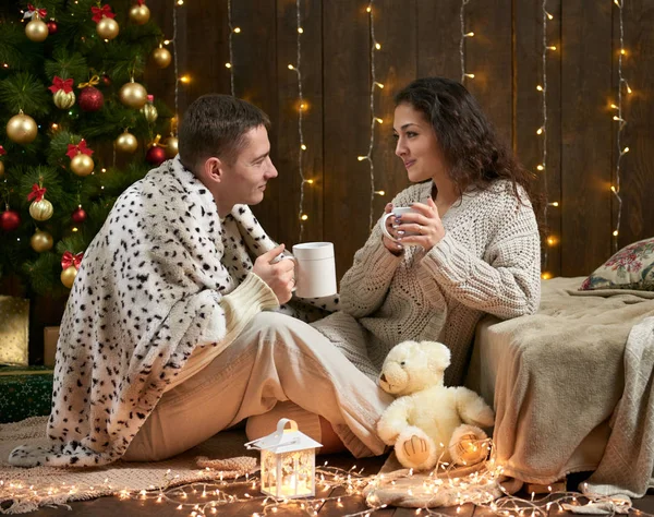 La coppia giovane beve il tè, si siede su pavimento in interno scuro di legno con luci. Serata romantica e concetto d'amore. Vacanza di anno nuovo. Luci e decorazioni natalizie. Vestita di bianco . — Foto Stock