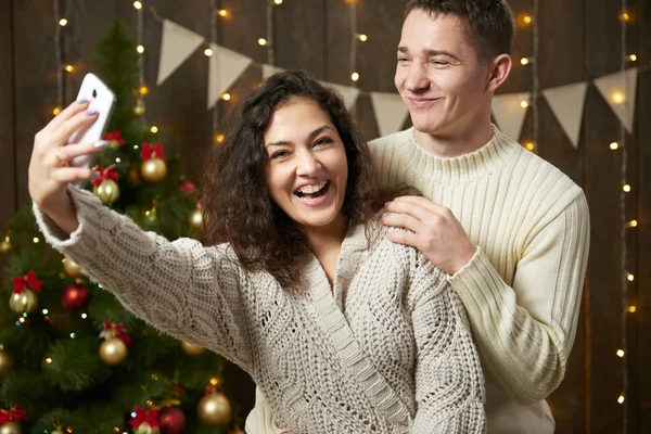 Coppia felice prendendo selfie e divertirsi nella decorazione di Natale. Interno in legno scuro con luci. Serata romantica e concetto d'amore. Vacanza di anno nuovo . — Foto Stock