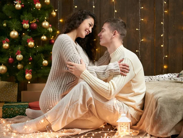 Молода пара сидить на підлозі в темному дерев'яному інтер'єрі з вогнями. Романтичний вечір і концепція любові. Новорічне свято. Різдвяні вогні та прикраси. одягнені в білий . — стокове фото