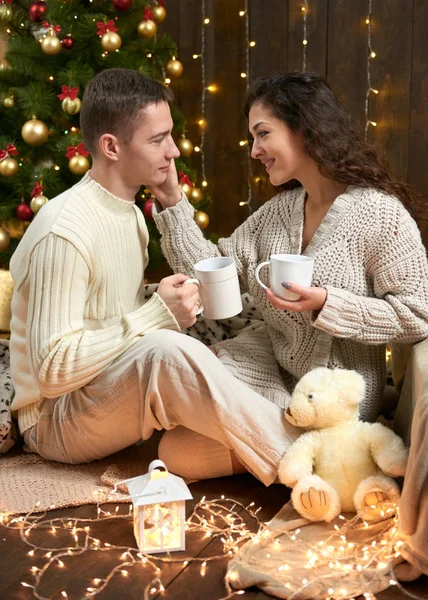 Coppia felice bere il tè e parlare nella decorazione natalizia, seduto sul pavimento in legno scuro interno con luci. Serata romantica e concetto d'amore. Vacanza di anno nuovo . — Foto Stock