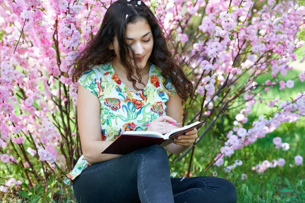 Portrait of young girl sitting near bush with pink flowers and read the book — Stock Photo, Image