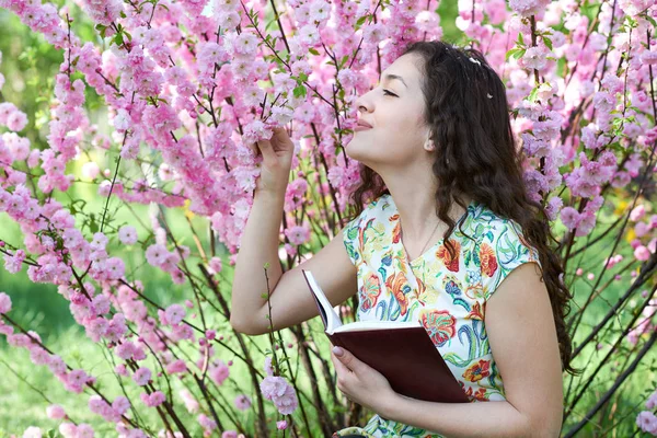 portrait of young girl sitting near bush with pink flowers and reading the book