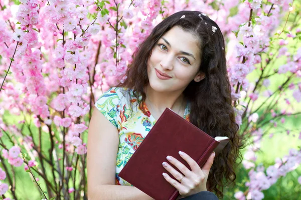 Portrait of young girl sitting near bush with pink flowers and reading the book — Stock Photo, Image