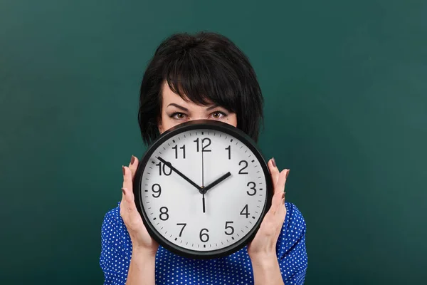 Mujer esconde su cara detrás del reloj, posando por pizarra, tiempo y concepto de educación, fondo verde, toma de estudio — Foto de Stock
