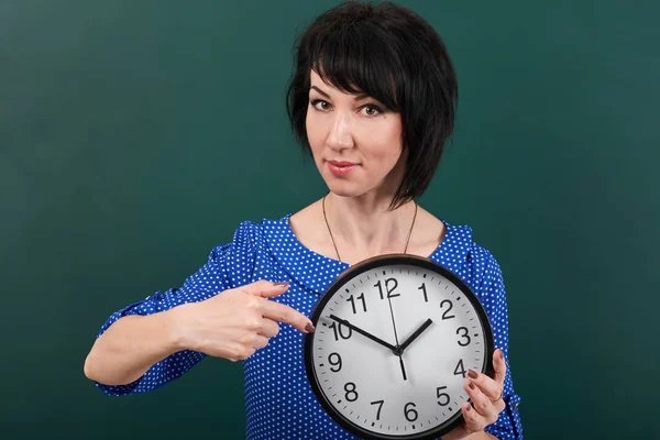 Mujer señalando con el dedo el reloj, posando por pizarra, tiempo y concepto de educación, fondo verde, toma de estudio — Foto de Stock