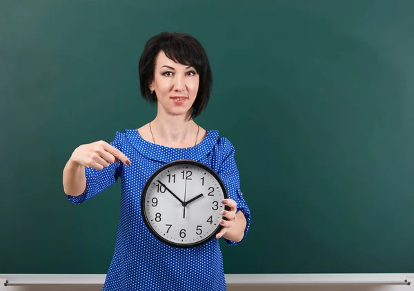 Frau zeigt mit dem Finger auf die Uhr, posiert mit Kreidetafel, Zeit- und Bildungskonzept, grüner Hintergrund, Studioaufnahme — Stockfoto