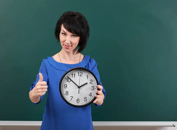 Woman with big clock shows Okey gesture, chalk board background, time and education concept, green background, studio shot — Stock Photo, Image