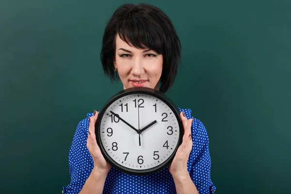 Woman hides her face behind the clock, posing by chalk board, time and education concept, green background, studio shot — Stock Photo, Image