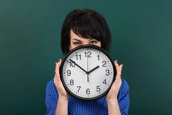 woman hides her face behind the clock, posing by chalk board, time and education concept, green background, studio shot