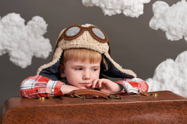 Мальчик, одетый как пилот самолета, сидит между облаками со старым чемоданом и снами
