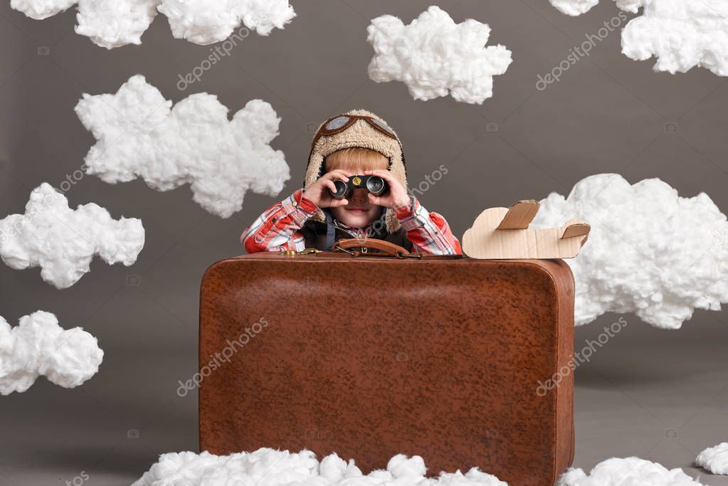boy dressed as an airplane pilot sit between the clouds with old suitcase and playing with handmade plane