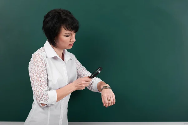 Une femme regarde à travers une loupe sur montre, un tableau noir comme arrière-plan, un concept d'éducation et d'affaires — Photo