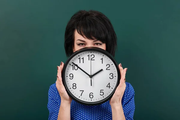 Woman hides her face behind the clock, posing by chalk board, time and education concept, green background, studio shot — Stock Photo, Image