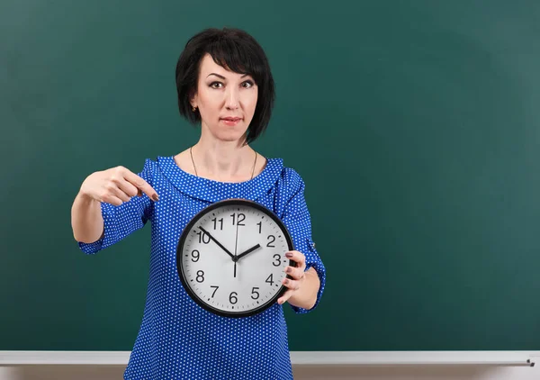 Frau zeigt mit dem Finger auf die Uhr, posiert mit Kreidetafel, Zeit- und Bildungskonzept, grüner Hintergrund, Studioaufnahme — Stockfoto