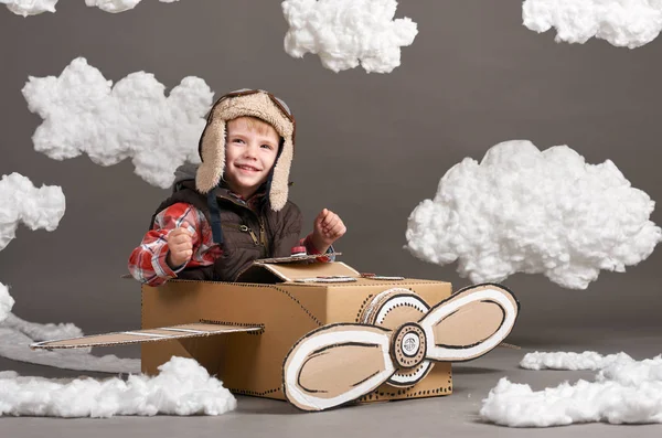Το αγόρι που παίζει σε ένα αεροπλάνο από χαρτόκουτο και ονειρεύεται να γίνει πιλότος, σύννεφα, βαμβάκι σε ένα γκρίζο φόντο — Φωτογραφία Αρχείου