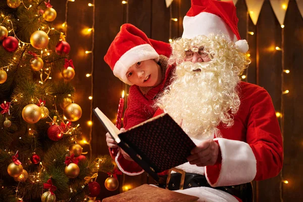 Père Noël et santa aide garçon livre de lecture, assis à l'intérieur près décoré arbre de Noël avec des lumières Joyeux Noël et Joyeuses Fêtes ! — Photo