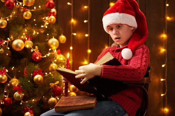 Adolescent garçon lecture livre, assis à l'intérieur près décoré arbre de Noël avec des lumières, habillé comme Père Noël helper Joyeux Noël et Joyeuses Fêtes ! — Photo