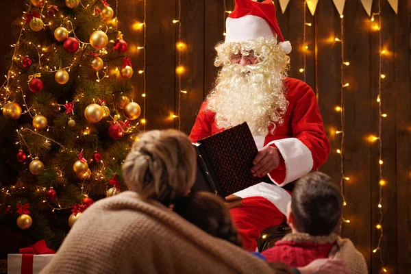 Père Noël livre de lecture pour la famille. Mère et enfants assis à l'intérieur près de l'arbre de Noël décoré avec des lumières Joyeux Noël et Joyeuses Fêtes ! — Photo