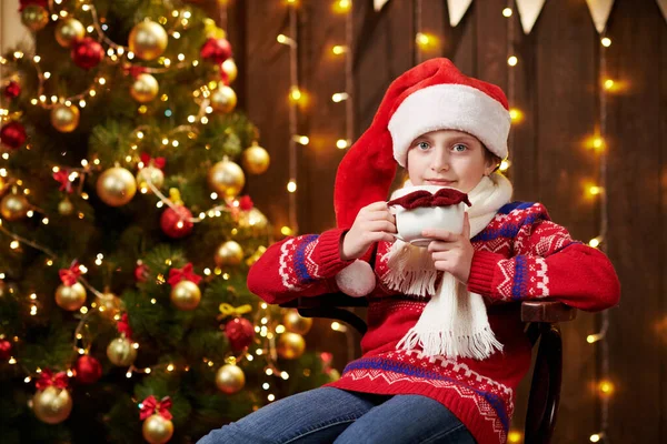 Веселе дівчисько з Санта-Хелпер з чашкою чаю сидить у приміщенні біля прикрашеного ксам дерева зі світлом, одягненим у червоний светр - Веселого Різдва та Веселих свят.! — стокове фото