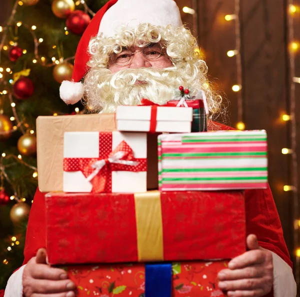 Père Noël posant avec de nombreux cadeaux, assis à l'intérieur près de l'arbre de Noël décoré avec des lumières Joyeux Noël et Joyeuses Fêtes ! — Photo