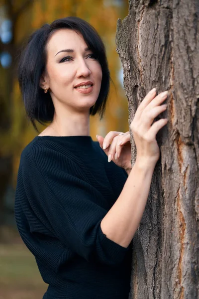 Schöne elegante Frau, die im Herbst in einem Stadtpark neben einem Baum steht und posiert — Stockfoto