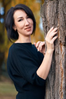 Güzel, zarif bir kadın. Sonbaharda bir şehir parkında ağaç kenarında durup poz veriyor.