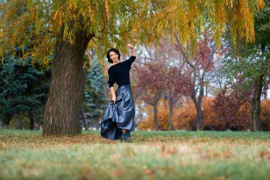 Sonbaharda şehir parkında büyük sarı bir ağacın yanında duran ve poz veren zarif bir kadın.