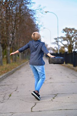 Sonbahar parkında yürüyen ve dönen mutlu sarışın kadın, kot pantolon ve ceket giymiş.