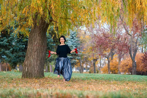 가을에 밝은 노란 나무들 이 도시 공원에서 붉은 구두를 손에 들고 맨발로 뛰고 있는 아름다운 여인 — 스톡 사진