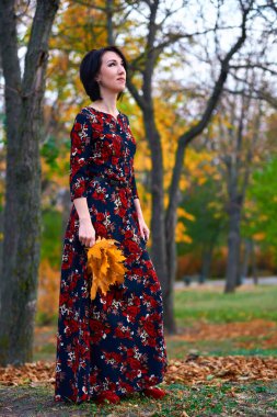 Güz parkında ayakta duran ve sarı yapraklarla poz veren zarif bir kadın.