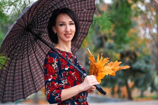 Schöne elegante Frau steht und posiert mit Strauß gelber Blätter und Regenschirm im herbstlichen Stadtpark — Stockfoto