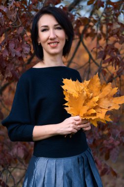 Güz parkında ayakta duran ve sarı yapraklarla poz veren zarif bir kadın.