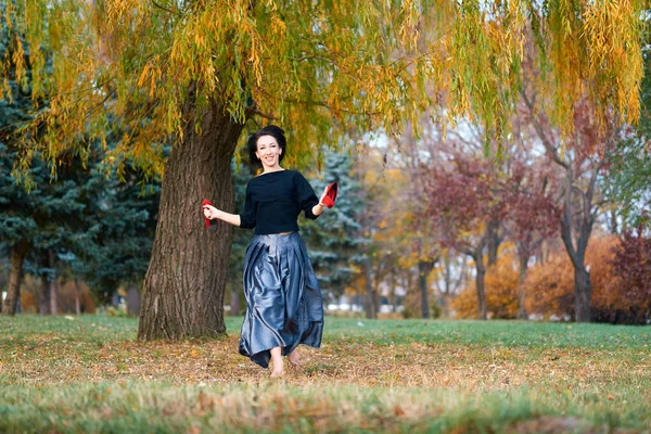 Hermosa mujer elegante corriendo descalza con zapatos rojos en la mano en un parque de la ciudad en otoño, árboles de color amarillo brillante —  Fotos de Stock