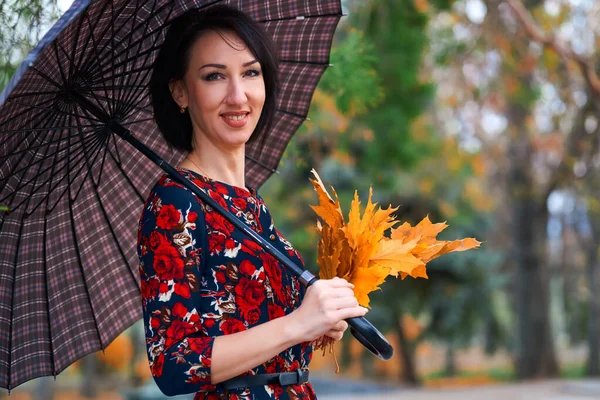 Piękna elegancka kobieta stojąca i pozująca z bukietem żółtych liści i parasolem w jesiennym parku miejskim — Zdjęcie stockowe