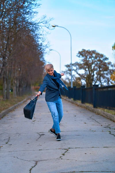 幸せなブロンドの女性歩くと秋の都市公園を回転,青いジーンズとジャケットを身に着けています — ストック写真