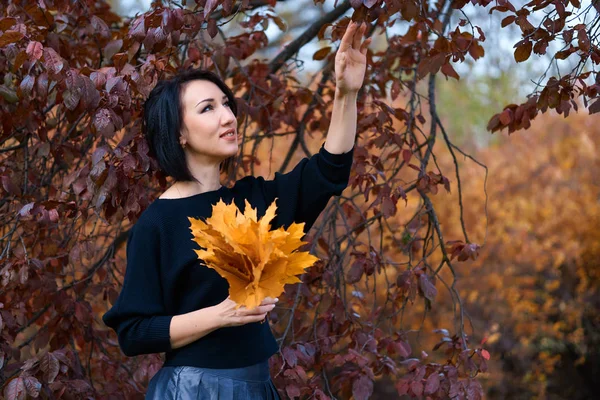 Krásná elegantní žena stojící a pózující s kyticí žlutých listů v podzimním městském parku — Stock fotografie