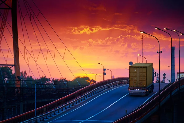 Φορτηγό με βόλτες με κοντέινερ πάνω από τη γέφυρα, όμορφο ηλιοβασίλεμα, φορτηγά αυτοκίνητα στο βιομηχανικό λιμάνι, ο δρόμος ανεβαίνει — Φωτογραφία Αρχείου