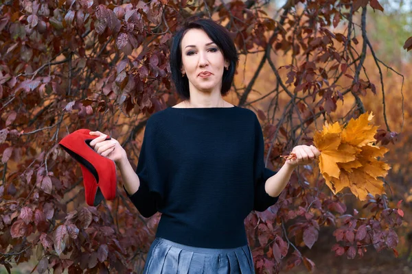 Mulher elegante bonita de pé e posando com buquê de folhas amarelas e sapatos vermelhos no parque da cidade de outono — Fotografia de Stock