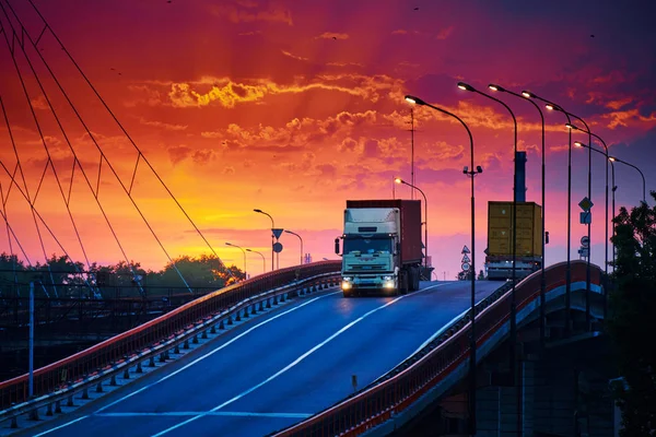 Truck with container rides over the bridge, beautiful sunset, freight cars in industrial seaport, the road goes up — Stock Photo, Image