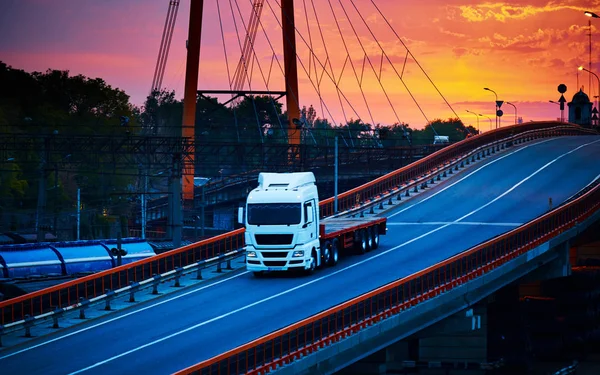 Truck with container rides over the bridge, beautiful sunset, freight cars in industrial seaport, the road goes up — Stock Photo, Image