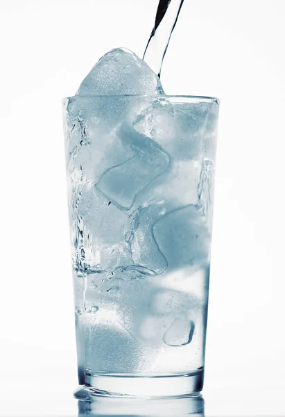 Наливая воду в стакан, полный кубиков льда, белый фон, синий тонированный объект — стоковое фото