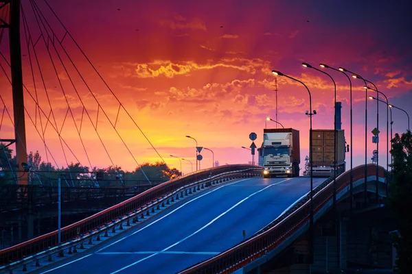 Truck with container rides over the bridge, beautiful sunset, freight cars in industrial seaport, the road goes up — Stock Photo, Image