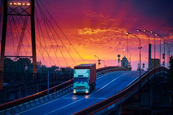Truck with container rides over the bridge, beautiful sunset, freight cars in industrial seaport, the road goes up — Stock Photo, Image