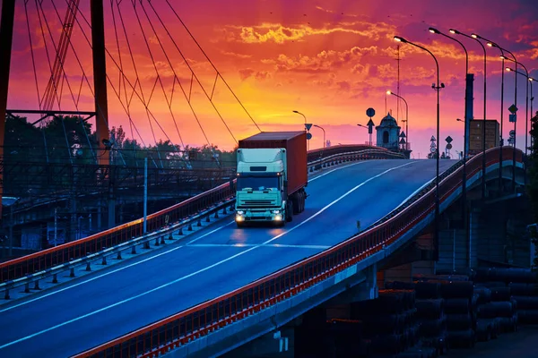 Truck with container rides over the bridge, beautiful sunset, freight cars in industrial seaport, the road goes up — Stock Photo, Image