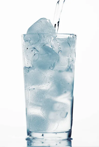 Наливая воду в стакан, полный кубиков льда, белый фон, синий тонированный объект — стоковое фото